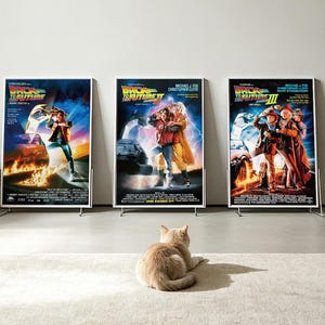 Peut inclure: Trois affiches de film encadrées pour Back to the Future, Back to the Future Part II et Back to the Future Part III, présentant chacune la voiture DeLorean et les personnages principaux, alignées avec un chat clair assis au premier plan.