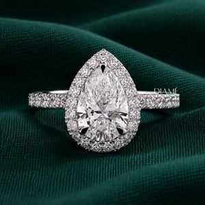 May include: A pear-shaped diamond ring featuring a central pear-cut stone surrounded by a halo of smaller round diamonds. The band is also encrusted with small round diamonds along the sides. The word DIAMÉ is printed in white on the right side.