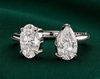 May include: A silver-toned open ring featuring two prominent clear stones, one in an oval cut and the other in a pear cut, each secured by four prongs. The band is smooth and polished, and the word DIAMÉ is printed in small white letters on the right.
