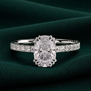 May include: A silver-toned ring featuring a large, oval-cut center stone held by multiple prongs. The band is adorned with a series of small, round-cut stones along the sides. The word DIAMÉ is printed in small white letters on the right side.
