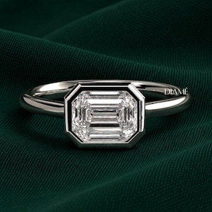 May include: A silver-toned ring featuring a central emerald-cut clear stone set in a smooth bezel setting. The band is polished and metallic. The word DIAMÉ is printed in small white letters on the right side of the ring setting.