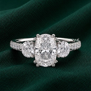 May include: A silver-toned ring featuring a large oval-cut center stone flanked by two smaller round-cut stones. The band is encrusted with small pavé-set stones along the sides. The word DIAMÉ is printed in small white letters on the right side.