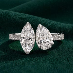 May include: A silver-toned ring featuring a large marquise-cut stone and a pear-cut stone set side-by-side. The band is encrusted with small pavé stones along the sides. The word DIAMÉ is printed in small white letters on the right side of the ring.