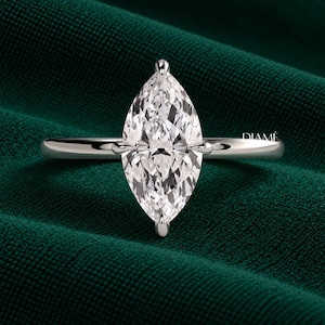 May include: A silver-toned ring featuring a large, faceted marquise-cut clear stone held by a six-prong setting on a smooth, polished band. The word DIAMÉ is printed in small white capital letters on the right side of the ring band.