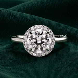 May include: A silver-toned ring featuring a large, round, faceted centre stone surrounded by a halo of smaller, round, brilliant-cut stones. The band is smooth and polished. The word DIAMÉ is printed in small white letters on the right side.