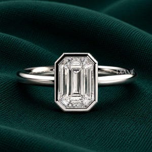 May include: A silver-toned ring featuring a large, rectangular emerald-cut clear gemstone set in a smooth bezel setting. The band is polished and metallic. The word DIAMÉ is printed in small white letters on the right side of the ring setting.