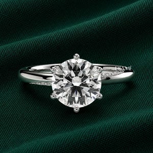 May include: A silver-toned ring featuring a large, round, faceted center stone held by six prongs. The band is smooth and polished, with a delicate row of small, sparkling stones embedded along the shoulders. The word DIAMÉ is visible.
