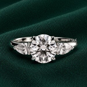 May include: A silver-toned ring featuring a large, round, brilliant-cut centre stone flanked by two smaller, pear-shaped stones on either side. The metal band is polished and smooth, and the word DIAME is visible on the right side of the setting.