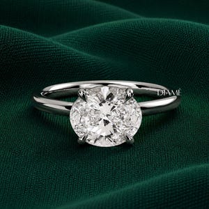 May include: A silver-toned ring featuring a large, oval-cut clear gemstone set in a four-prong setting. The band is smooth and polished, with the brand name DIAMÉ printed in small white letters on the right side of the ring setting.