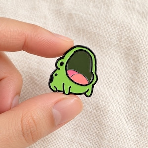 Op de afbeelding: Een kleine emaille speld in de vorm van een cartoonkikker met een felgroene huid, een wijd open donkergroene mond en een zichtbare roze tong. De kikker heeft zwarte omlijningen, twee kleine zwarte ogen en vier korte ledematen in een platte stijl.