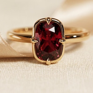 May include: A gold ring featuring a large, rectangular cushion-cut deep red gemstone set in a four-prong mounting. The band is smooth and polished, showcasing a classic design with a vibrant, faceted center stone that reflects light.