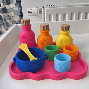 Puede incluir: Un juego de juguetes de silicona para hacer pociones sobre una bandeja rosa con forma de nube. Incluye tres botellas con tapones marrones, un caldero azul con una cuchara amarilla y dos vasos pequeños con iconos en relieve.