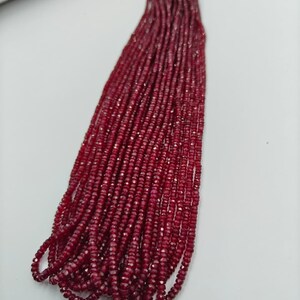 May include: A long strand of small, faceted, deep red ruby beads arranged in a vertical cluster. The beads are uniform in size and shape, featuring a reflective, polished surface that catches the light, creating a dense, textured appearance.