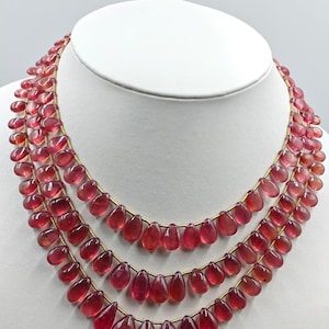 May include: A multi-strand necklace featuring three layers of teardrop-shaped, translucent pinkish-red gemstone beads strung on thin gold-toned wire. The beads are uniform in shape and size, creating a cascading effect around the neck.