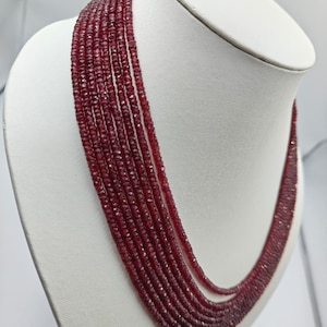 May include: A multi-strand necklace featuring seven rows of small, faceted, deep red gemstone beads. The beads are uniform in size and shape, creating a layered effect that drapes around the neck. The deep red stones have a subtle sparkle.