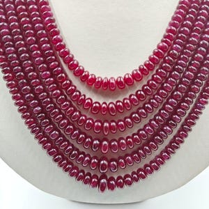 Puede incluir: Un collar de múltiples hilos con cinco capas de cuentas de rubí de color rojo intenso, lisas y redondas. Las cuentas son uniformes en forma y tamaño, dispuestas en un diseño en cascada que crea una exhibición densa de esferas pulidas.
