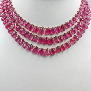 Può includere: Una collana a tre fili composta da numerose perle traslucide a forma di goccia di colore rosa intenso, infilate su un sottile filo dorato. Le perle sono disposte in tre strati a cascata, creando un design uniforme e simmetrico.