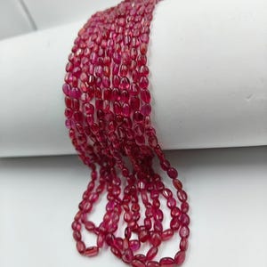 Könnte beinhalten: Eine Ansammlung mehrerer Stränge aus kleinen, ovalen, durchscheinenden, tief rosa-roten Perlen. Die Perlen sind in langen, vertikalen Linien aufgereiht und bilden ein dichtes, gleichmäßiges Bündel aus polierten, reflektierenden Elementen.