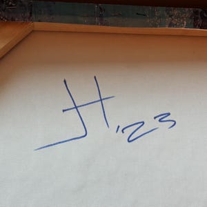 May include: A light-colored canvas stretched over a wooden frame, featuring a blue handwritten signature that reads H '23. The texture of the woven fabric is visible, and the wooden stretcher bars are partially visible at the top edge of the canvas.