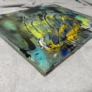 May include: A square canvas painting featuring a layered abstract composition with vibrant splashes of yellow, deep blue, and teal paint. The surface displays textured black splatters, dark streaks, and glossy highlights that create depth across the piece.