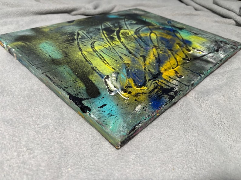May include: A square canvas painting featuring a layered abstract composition with vibrant splashes of yellow, deep blue, and teal paint. The surface displays textured black splatters, dark streaks, and glossy highlights that create depth across the piece.