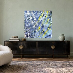 May include: A square abstract painting featuring a textured composition of deep blue, light blue, and white paint strokes, accented by vibrant yellow streaks. The artwork displays a layered, energetic application of color across the entire canvas.