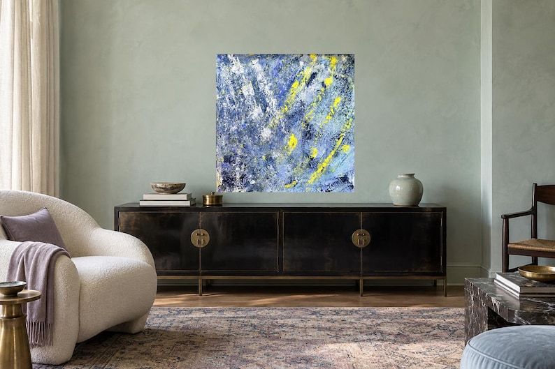 May include: A square abstract painting featuring a textured composition of deep blue, light blue, and white paint strokes, accented by vibrant yellow streaks. The artwork displays a layered, energetic application of color across the entire canvas.