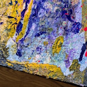 May include: An abstract painting on canvas featuring a thick, textured application of paint in shades of deep blue, bright yellow, light purple, and metallic gold, accented with vibrant red splatters across the surface of the artwork.
