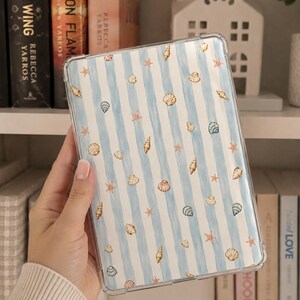 May include: A clear protective tablet case featuring a pattern of light blue vertical watercolor stripes interspersed with small illustrations of various seashells and orange starfish scattered across the surface of the cover.