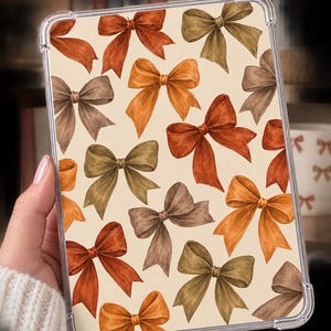 May include: A clear tablet case featuring a repeating pattern of watercolour-style bows in shades of burnt orange, olive green, and muted brown against a cream-coloured background. The case has reinforced corners and a transparent, flexible frame.