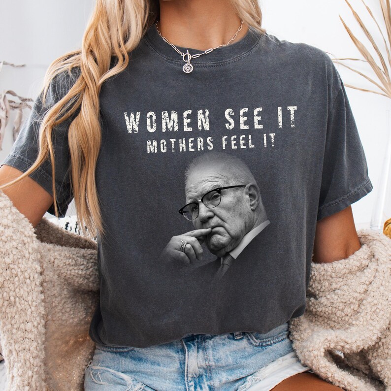 May include: A charcoal gray short-sleeve t-shirt featuring a black and white photographic print of a person wearing glasses with their hand near their chin. The text "WOMEN SEE IT MOTHERS FEEL IT" is printed in white above the portrait.