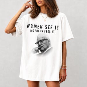May include: A white short-sleeved t-shirt featuring a black and white photographic print of a man wearing glasses with his hand near his chin. Above the image, the text reads "WOMEN SEE IT" and "MOTHERS FEEL IT" in a bold, distressed black font.
