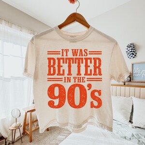 May include: A cream-colored short-sleeve t-shirt featuring the orange distressed text "IT WAS BETTER IN THE 90's" printed on the front. The graphic includes horizontal lines above and below the text, with the year "90's" in a large font.