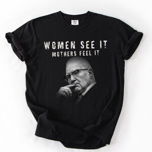 May include: A black short-sleeved t-shirt featuring a white graphic print of an older person wearing glasses with a hand near their chin. The text "WOMEN SEE IT MOTHERS FEEL IT" is printed in white capital letters above the portrait on the front of the shirt.