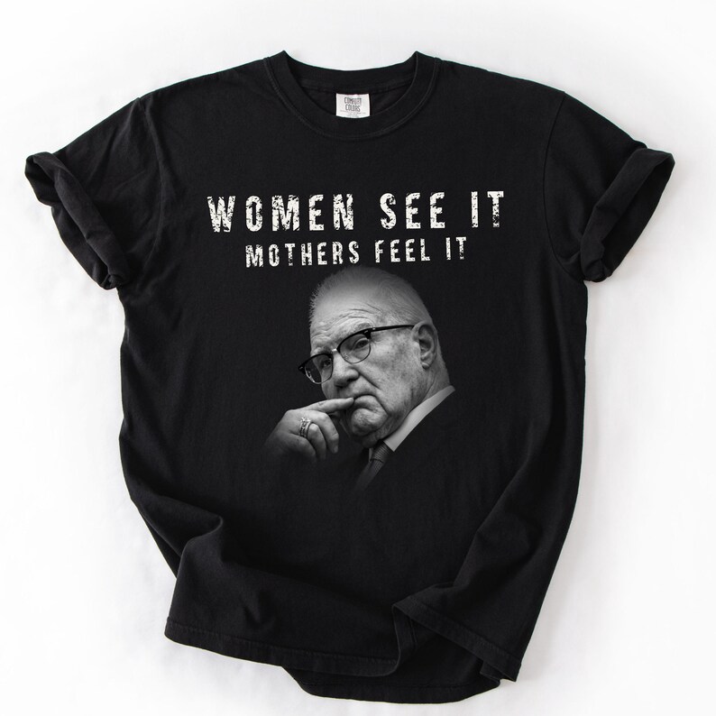 May include: A black short-sleeved t-shirt featuring a white graphic print of an older person wearing glasses with a hand near their chin. The text "WOMEN SEE IT MOTHERS FEEL IT" is printed in white capital letters above the portrait on the front of the shirt.