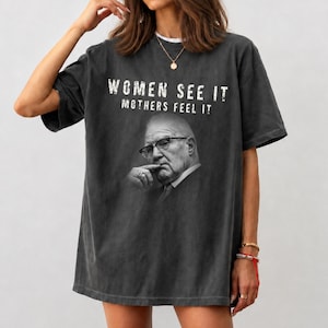 May include: A charcoal gray oversized t-shirt featuring a black and white photographic print of an older person wearing glasses with a hand near their chin. The text "WOMEN SEE IT MOTHERS FEEL IT" is printed in white distressed lettering above the portrait.