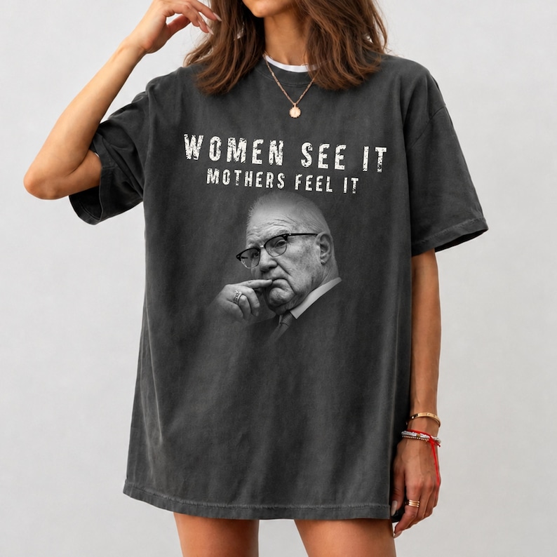 May include: A charcoal gray oversized t-shirt featuring a black and white photographic print of an older person wearing glasses with a hand near their chin. The text "WOMEN SEE IT MOTHERS FEEL IT" is printed in white distressed lettering above the portrait.