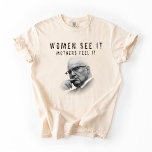 May include: A cream-colored short-sleeve t-shirt featuring a black and white portrait of an older person wearing glasses with a hand near their chin. The text "WOMEN SEE IT MOTHERS FEEL IT" is printed in a distressed black font above the portrait.