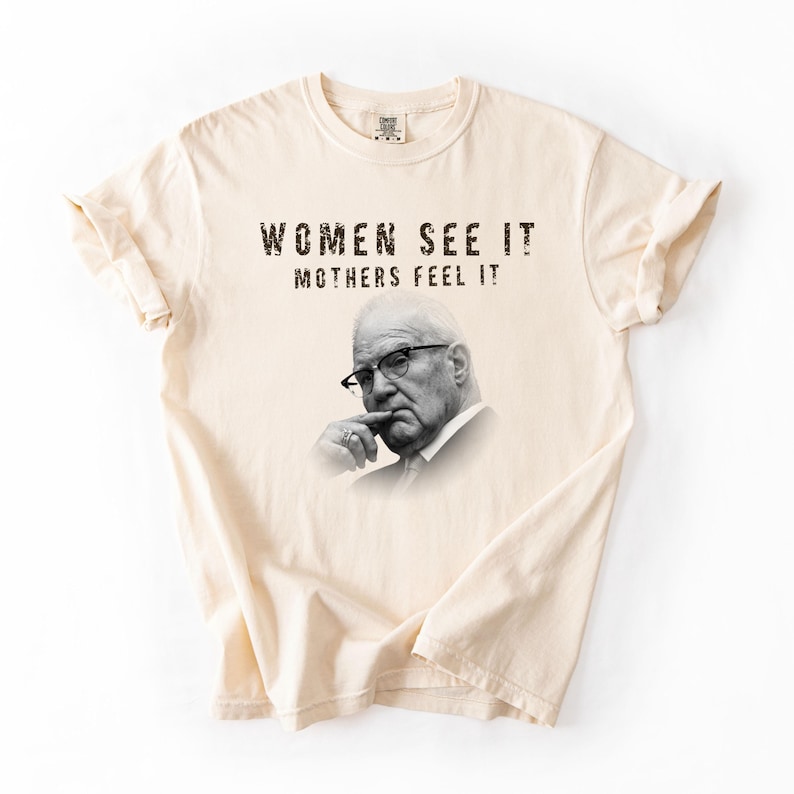 May include: A cream-colored short-sleeve t-shirt featuring a black and white portrait of an older person wearing glasses with a hand near their chin. The text "WOMEN SEE IT MOTHERS FEEL IT" is printed in a distressed black font above the portrait.
