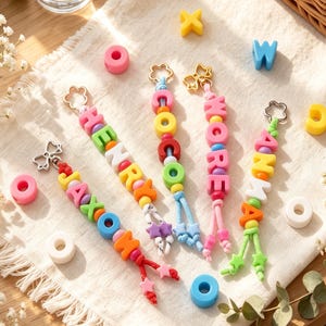 Op de afbeelding: Vijf kleurrijke kralen-sleutelhangers met letterkralen die de namen JAXON, HENRY, COCO, WORE en ANNA spellen. Elke sleutelhanger heeft een metalen bloemvormige sluiting, diverse ronde en stervormige kralen in felle kleuren en geknoopte koorduiteinden.