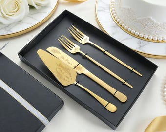 Könnte beinhalten: Ein goldfarbenes Torten-Servierset bestehend aus zwei Gabeln, einem gezackten Messer und einem Tortenheber, präsentiert in einer schwarzen Box. Messer und Heber sind mit floralen Gravuren sowie dem Text "The Troy's" und "Molly & Mike" versehen.