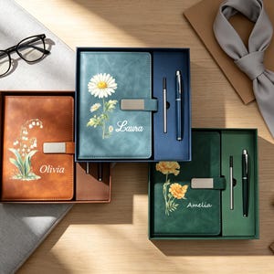 May include: Three rectangular gift boxes containing notebooks and pens. The brown notebook features a lily of the valley and the name Olivia. The blue notebook shows a daisy and the name Laura. The green notebook displays marigolds and the name Amelia.