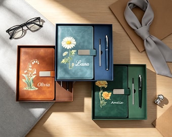 Könnte beinhalten: Drei rechteckige Geschenkboxen mit Notizbüchern und Stiften. Das braune Notizbuch zeigt ein Maiglöckchen und den Namen Olivia. Das blaue Notizbuch zeigt ein Gänseblümchen und den Namen Laura. Das grüne Notizbuch zeigt Ringelblumen und den Namen Amelia.