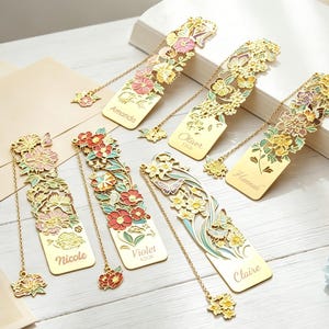 Puede incluir: Un conjunto de seis marcapáginas de metal dorado con intrincados recortes de flores y mariposas con detalles en esmalte rosa, rojo, amarillo y verde. Cada marcapáginas incluye un dije de cadena colgante y está grabado con los nombres Amanda, Oliver, Hannah, Nicole, Violet y Claire.