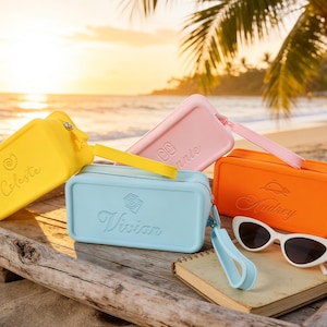 May include: Four rectangular silicone pouches in yellow, light blue, pink, and orange, each featuring a wrist strap, a zipper, and a personalized name embossed on the front. A pair of white cat-eye sunglasses rests beside the orange pouch.