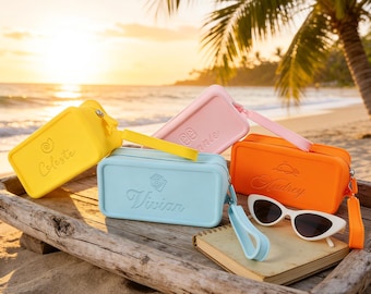 May include: Four rectangular silicone pouches in yellow, light blue, pink, and orange, each featuring a wrist strap, a zipper, and a personalized name embossed on the front. A pair of white cat-eye sunglasses rests beside the orange pouch.