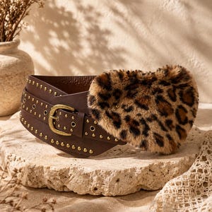 Può includere: Una cintura larga marrone con fibbia color ottone e file di piccoli rivetti metallici dorati. La cintura è decorata con una grande applicazione a forma di cuore in pelliccia sintetica con stampa leopardata marrone e nera sul davanti.