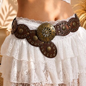 May include: A brown leather belt featuring circular segments adorned with brass-toned metal grommets and small studs. The center of the belt showcases a large, ornate, circular brass-toned buckle with intricate cut-out patterns and radial detailing.