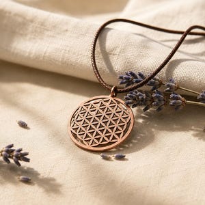 May include: A circular copper-toned pendant featuring a geometric flower of life pattern, suspended from a dark brown cord. The pendant is positioned alongside sprigs of dried purple lavender flowers on a textured off-white fabric surface.