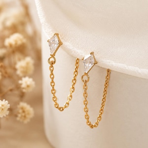 Pode incluir: Um par de brincos dourados com um pino em forma de diamante com uma pedra transparente, ligado a uma delicada corrente dourada que volta ao pino. O design cria um efeito pendente com um acabamento de metal polido e pedra facetada.